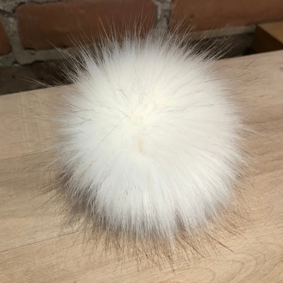 A fluffy white faux fur pom-pom ball resting on a light wood tabletop with a brick wall in the background.