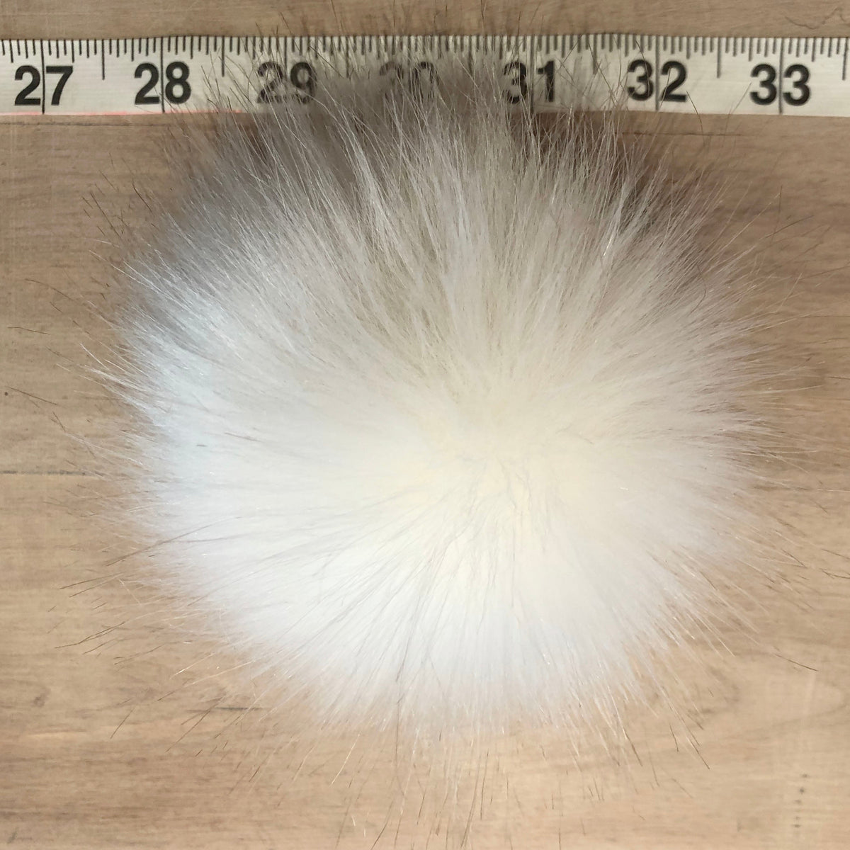 A fluffy white faux fur pom-pom ball shown on a wooden surface with a measuring tape ruler at the top for scale.
