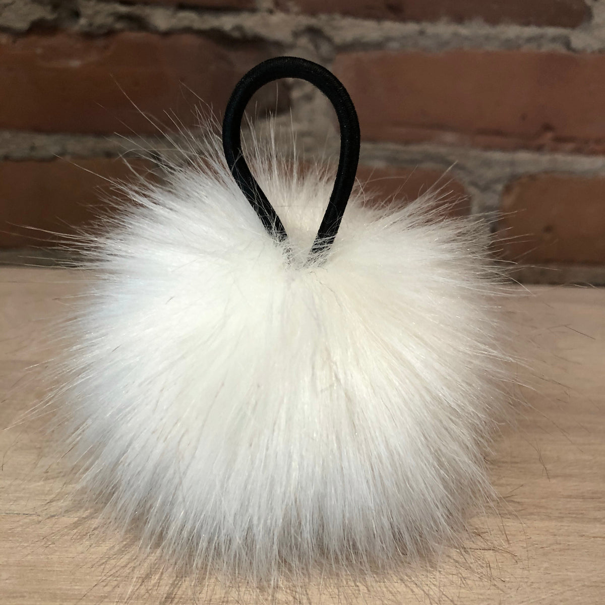 A fluffy white faux fur pom-pom keychain or bag charm with a black elastic loop, resting on a light wood surface with a brick wall background.