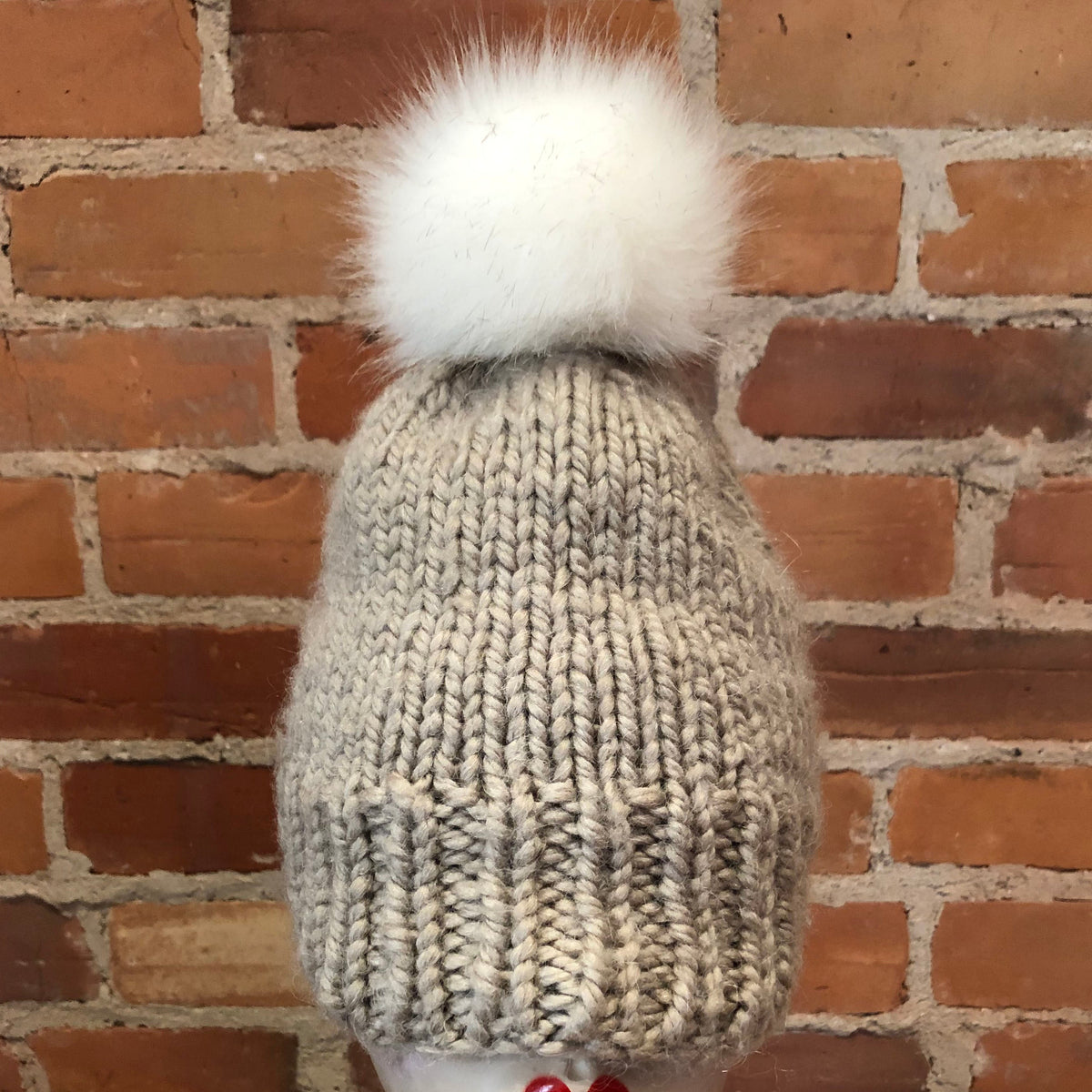 A light gray chunky knit beanie with a fluffy white pom-pom, displayed on a mannequin head against a red brick wall background.