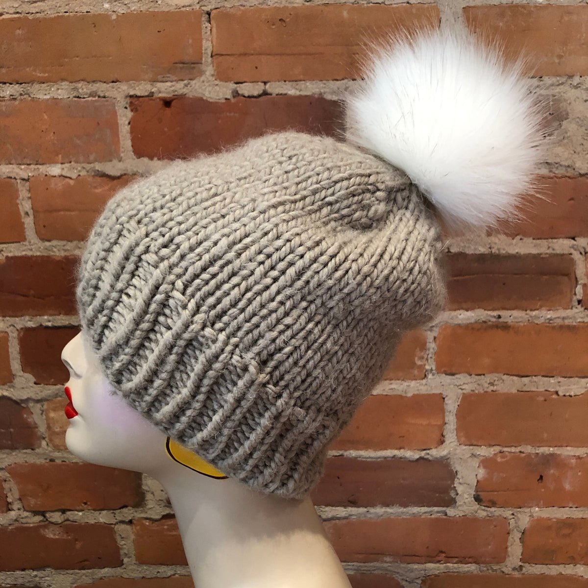 A light gray ribbed knit beanie with a large white faux fur pom-pom, displayed on a mannequin head against a red-brown brick wall background.