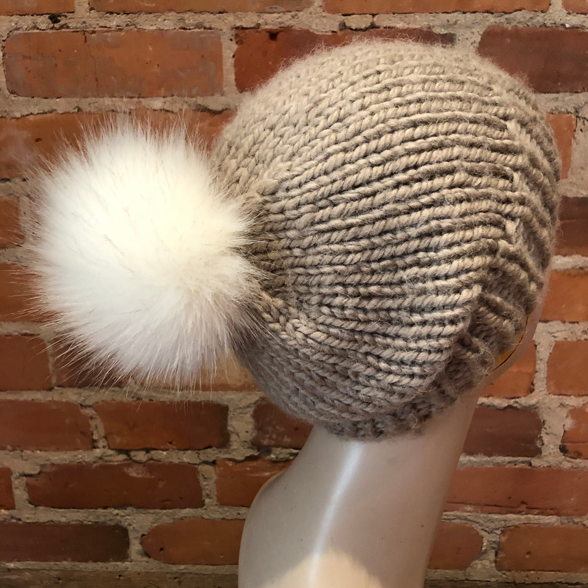 A chunky knit beanie hat in a warm taupe-beige color, featuring a large fluffy white faux-fur pom-pom, displayed on a mannequin head against a red brick wall background.