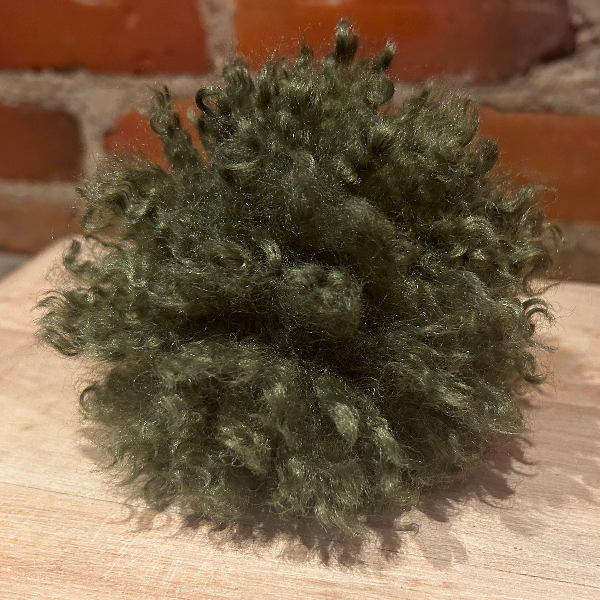 An olive green curly faux fur pom-pom toy resting on a light wooden surface with a blurred red brick wall in the background.
