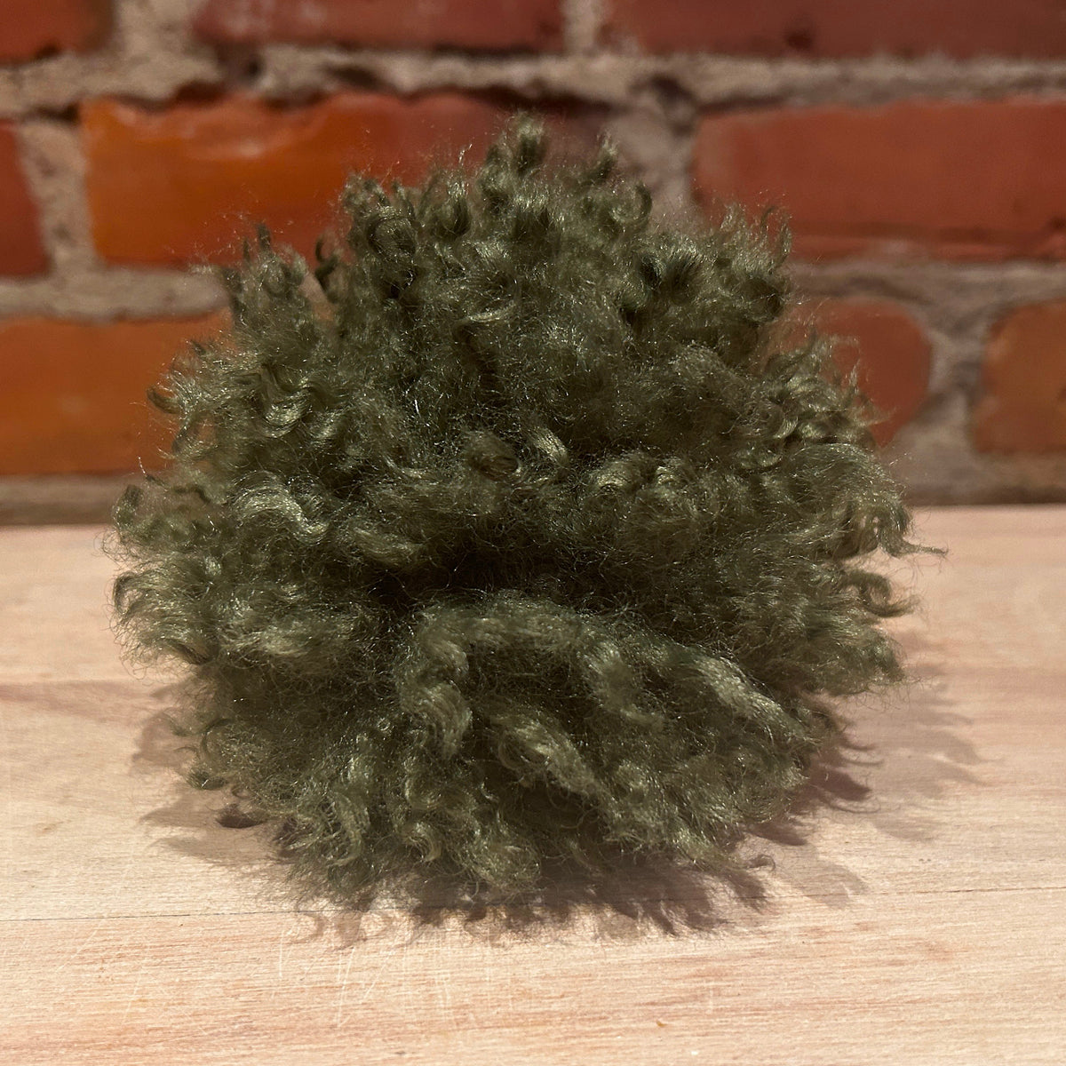 A small, fluffy, mossy green hat pom pom sits on a light-colored wooden surface with a blurred red brick wall background.