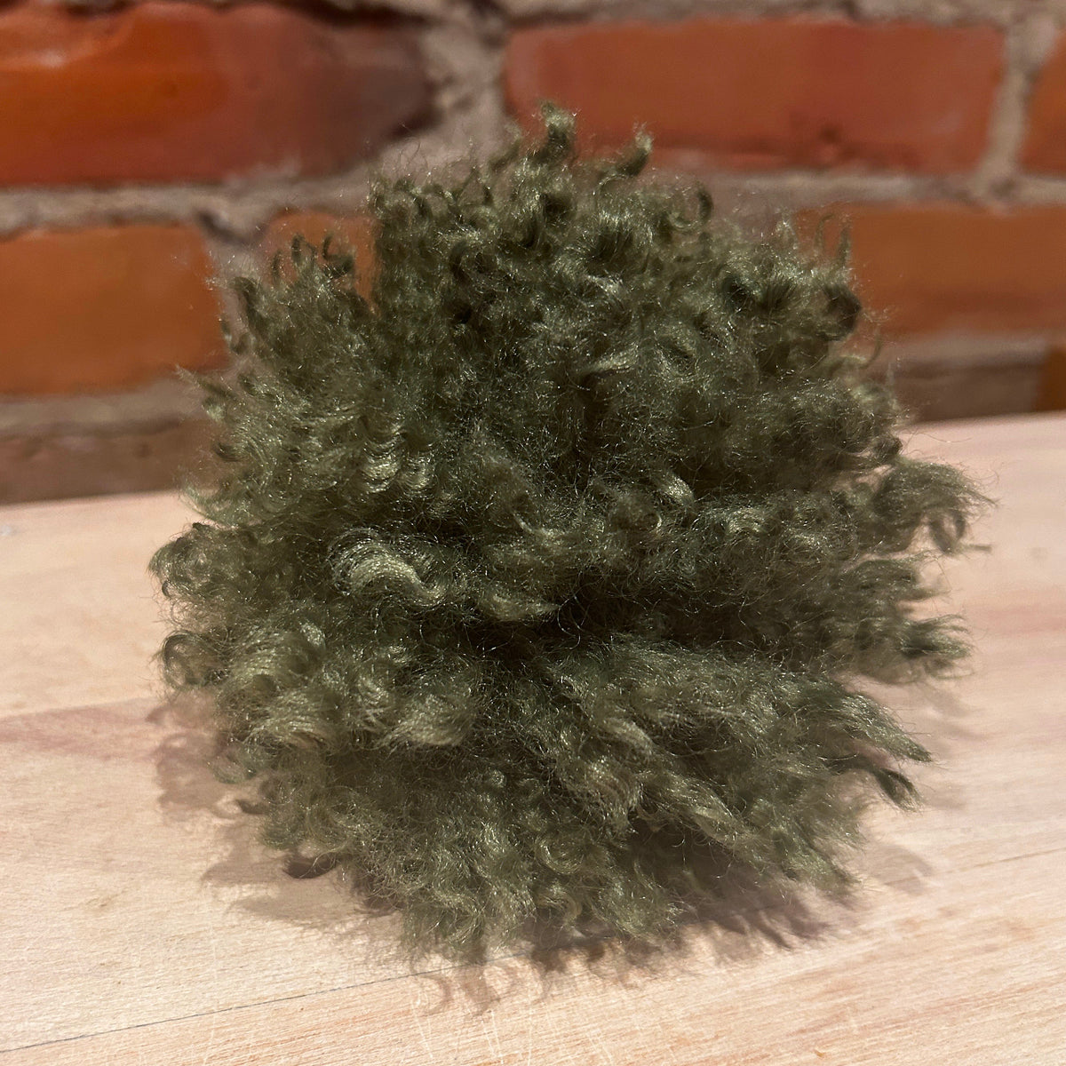 A small, fluffy, mossy green hat pom pom sitting on a light-colored wooden surface with a blurred red brick wall in the background.