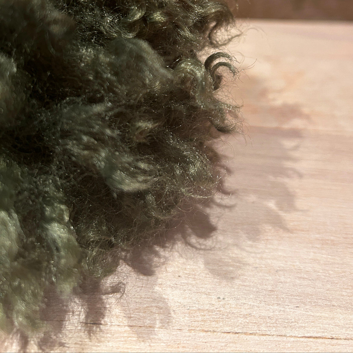 A close-up of a dark olive green curly pom pom, resting on a light wooden surface with a blurred red brick wall in the background.