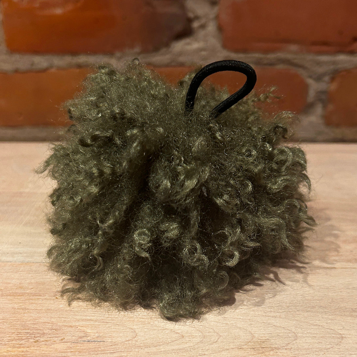 An olive green curly hat pom with a black elastic loop, resting on a light wood surface with a brick wall background.