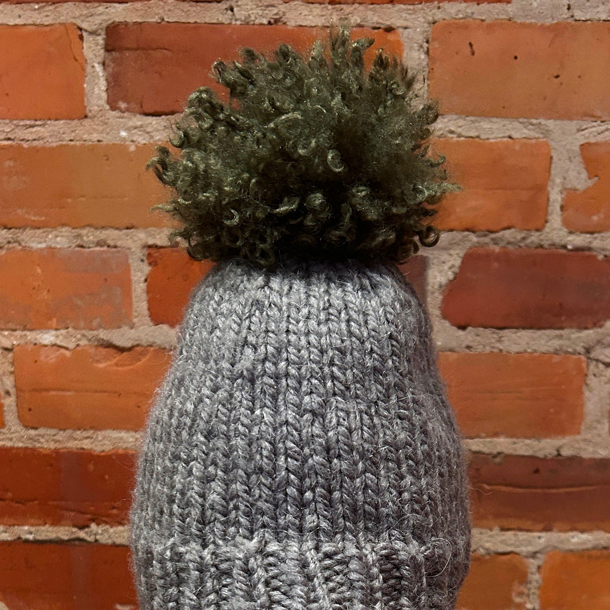 A gray ribbed knit beanie with a dark olive green pom-pom, displayed on a mannequin head with red lips in front of a red brick wall.