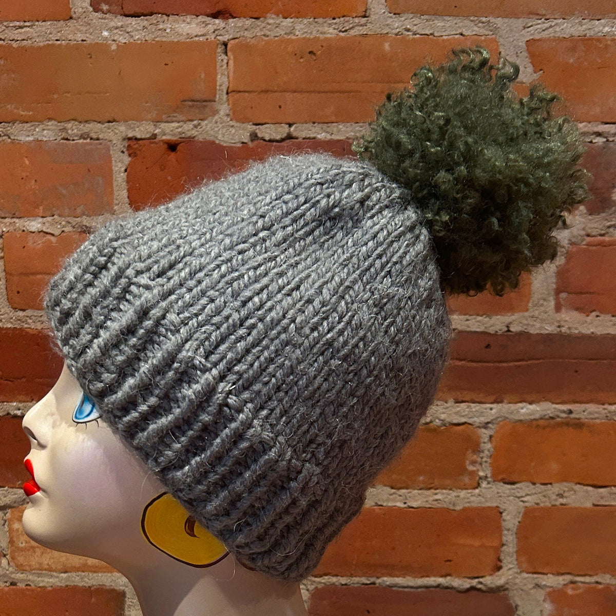 A gray ribbed knit beanie with a dark green pom-pom, displayed on a mannequin head against a red brick wall background.