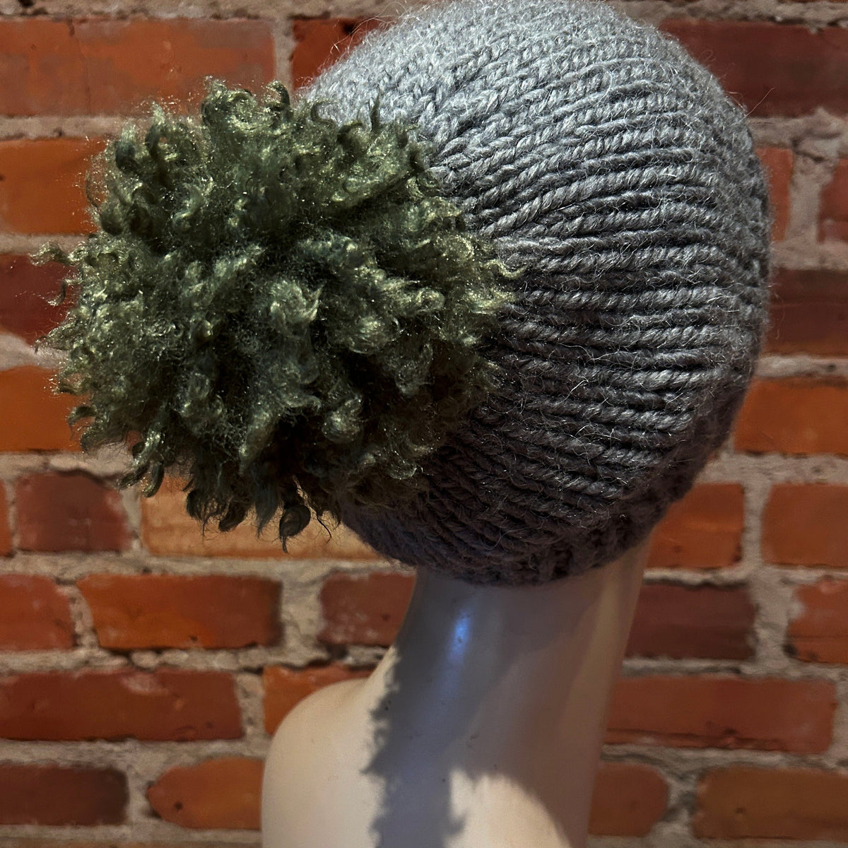 A gray ribbed knit beanie with a large textured green pom-pom, displayed on a mannequin head against a red brick wall background.