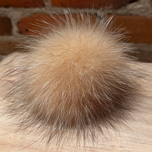 A close-up of a fluffy pom-pom ball made from light tan and cream fox fur, resting on a light wood surface with a blurred brick background.