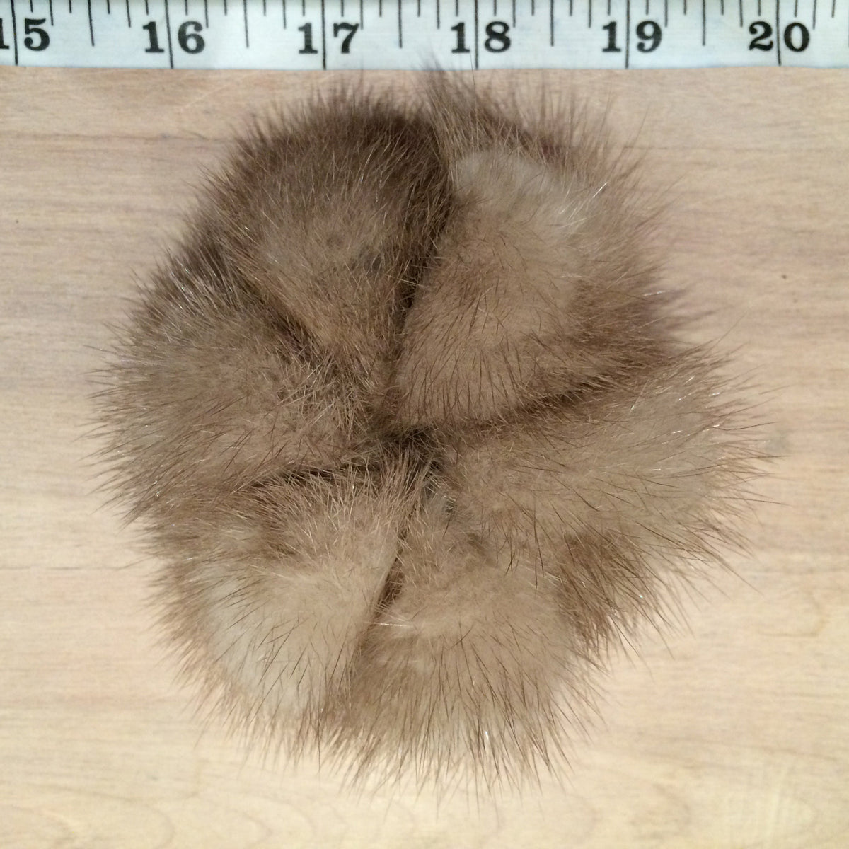 A brown mink fur pom-pom hair scrunchie laid flat on a light wood surface with a measuring tape ruler visible at the top for scale.