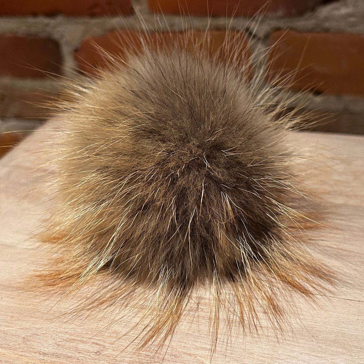 A close-up of a fluffy fox fur pom-pom in a natural brown and beige color, resting on a light wood surface with a blurred brick wall background.