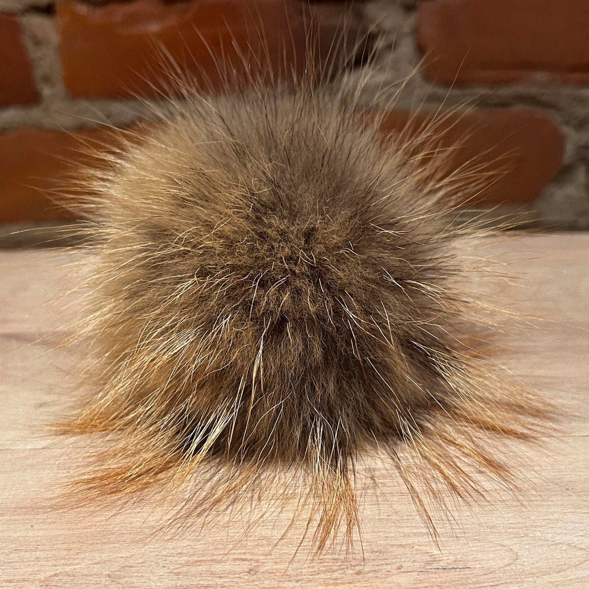 A round, fluffy fox fur pom-pom in shades of brown and beige, resting on a light wood surface with a blurred brick wall background.