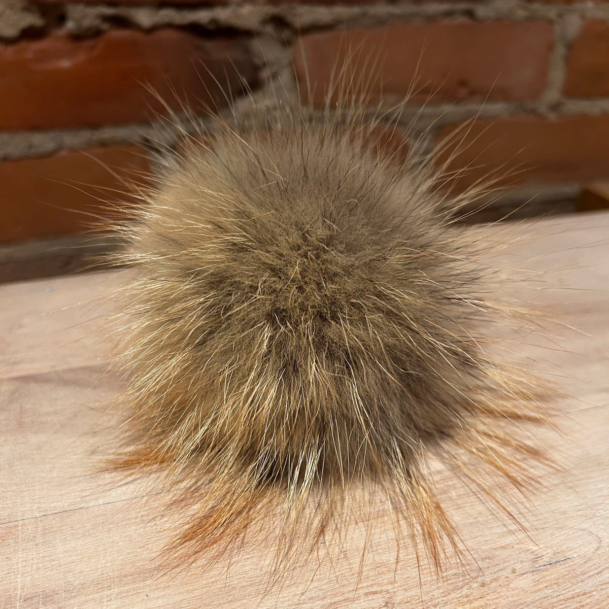 A fluffy pom-pom ball made from natural brown and beige animal fur, resting on a light wood surface with a brick wall in the background.