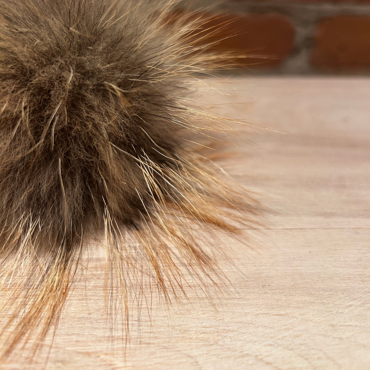 A close-up of a fluffy fox fur pom-pom accessory in brown and beige tones, resting on a light wood surface with a blurred brick wall background.