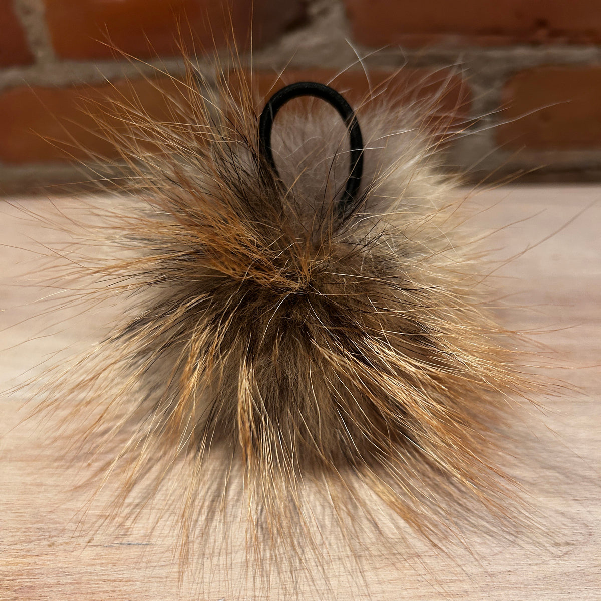 A fluffy fox fur pom-pom in natural brown and beige tones, featuring a black elastic loop at the top, resting on a light wood surface with a brick wall background.