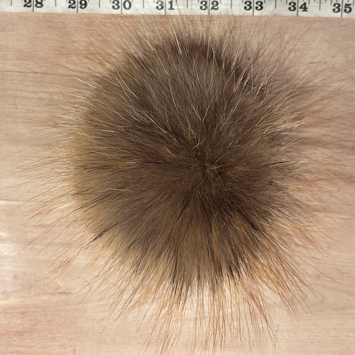 A round, fluffy fox fur pom-pom in a mix of brown and beige tones, shown on a light wood background with a measuring tape visible at the top edge.
