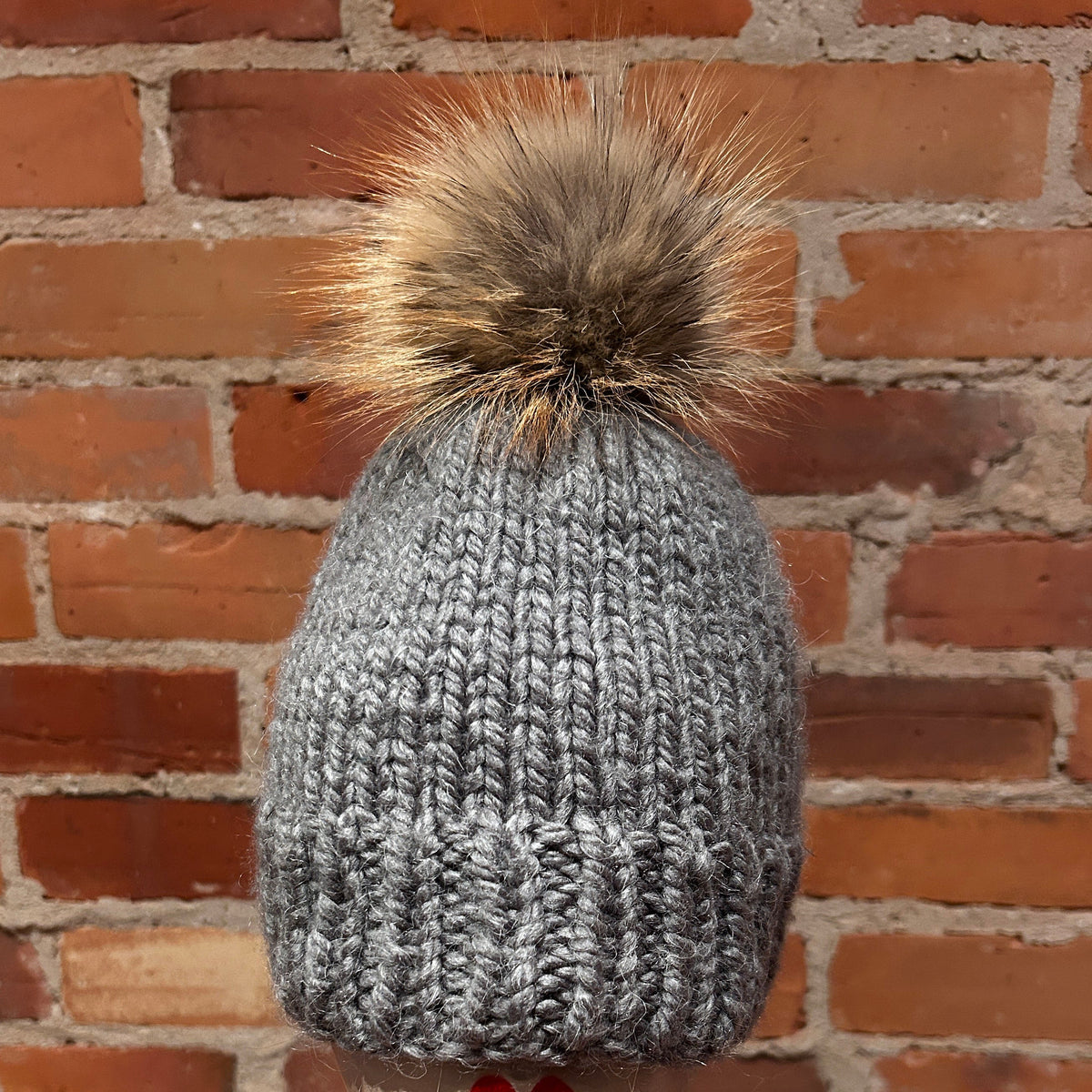 A gray ribbed knit beanie with a brown and beige fox fur pom-pom, displayed on a mannequin head with red lips in front of a red brick wall.