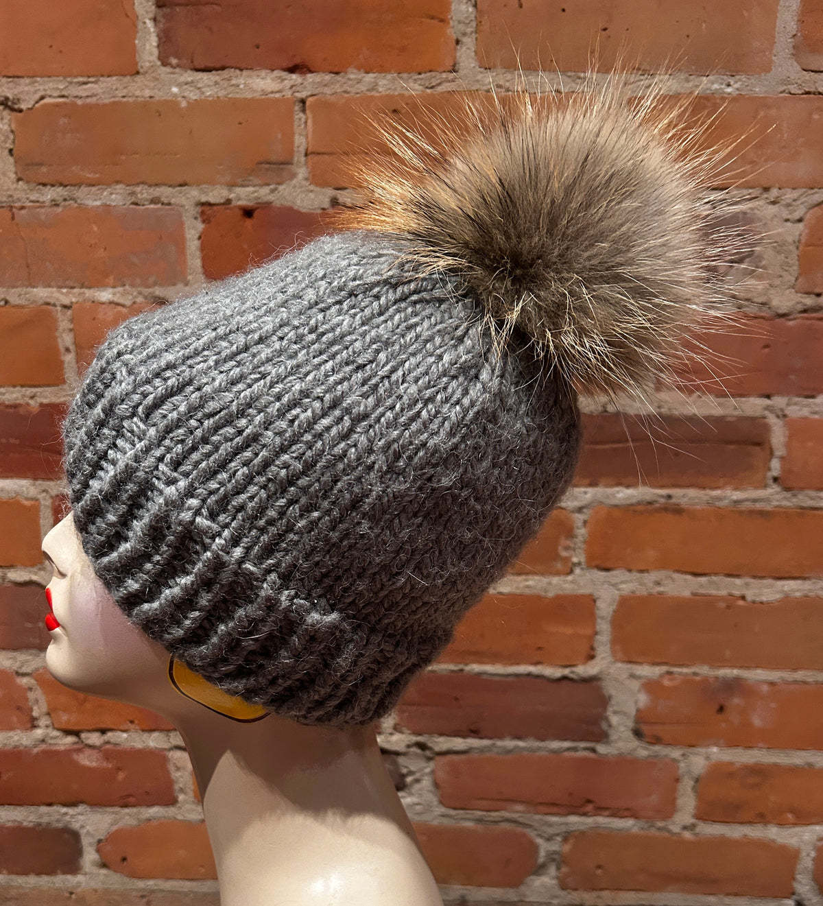 A gray ribbed knit beanie with a large brown and beige fox fur pom-pom, displayed on a mannequin head against a red brick wall background.