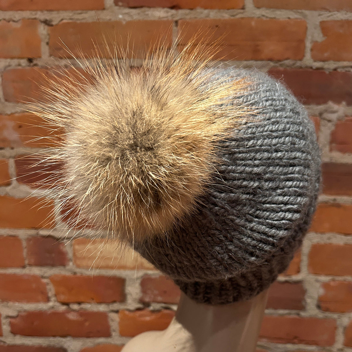 A gray knit beanie with a fluffy tan and brown fox -fur pom-pom, shown on a mannequin head against a red brick wall background.