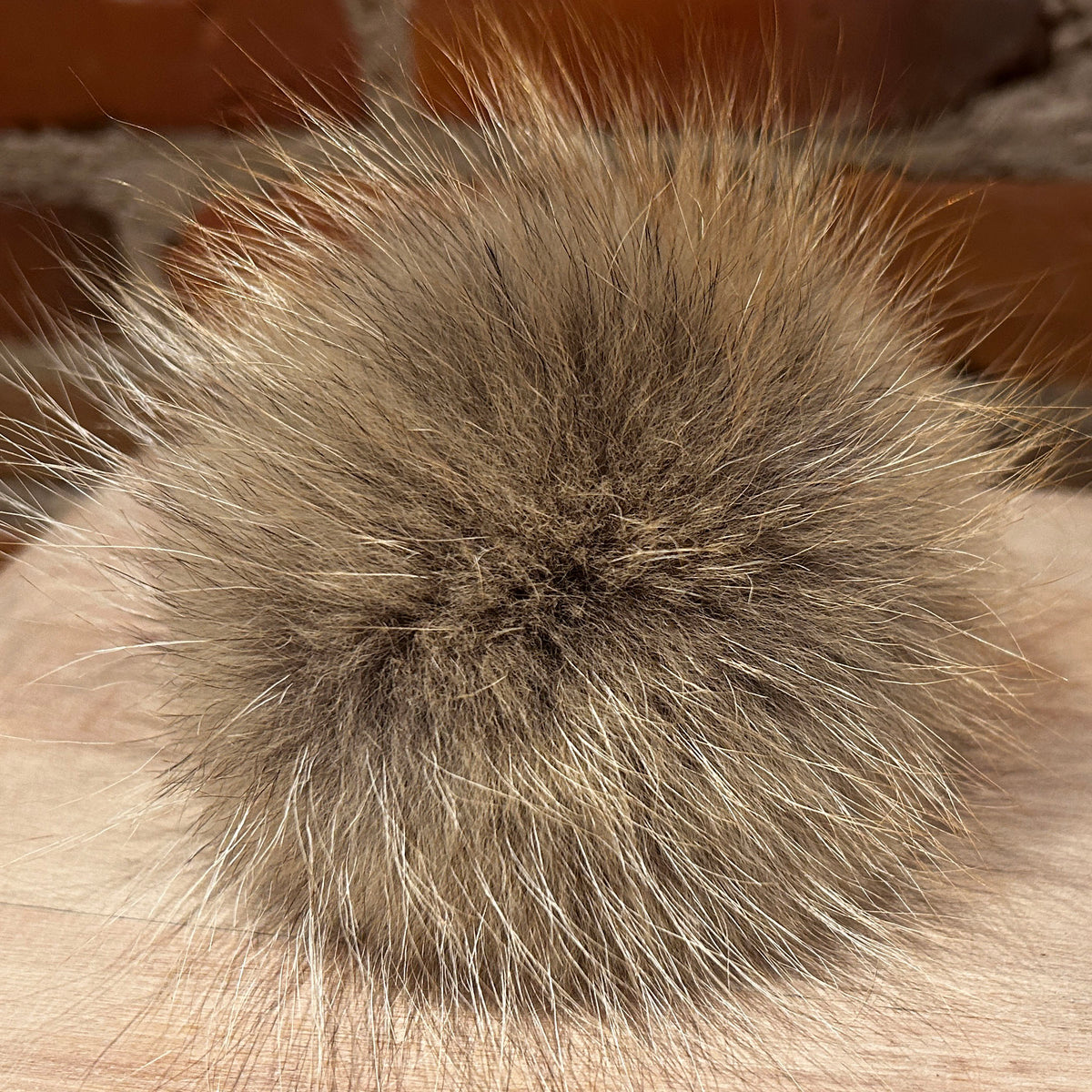 A close-up of a fluffy pom-pom made from natural brown and beige animal fur, resting on a light fabric surface with a blurred brick background.
