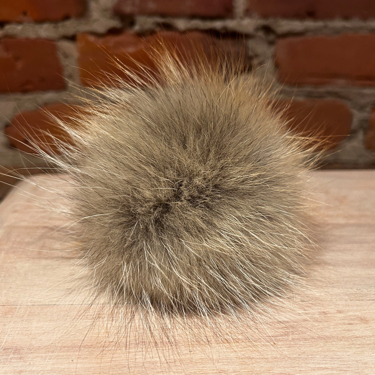 A fluffy Coyote-fur pom-pom in a warm taupe-brown color, resting on a light wood surface with a blurred red brick wall background.