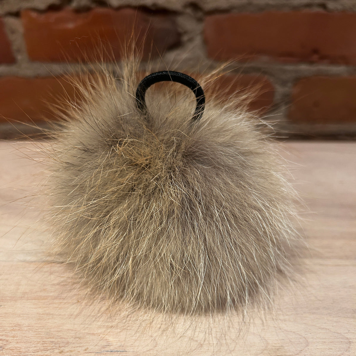 A fluffy Coyote-fur pom-pom in light brown and beige tones, attached to a black elastic loop, resting on a light wood surface with a brick wall background.