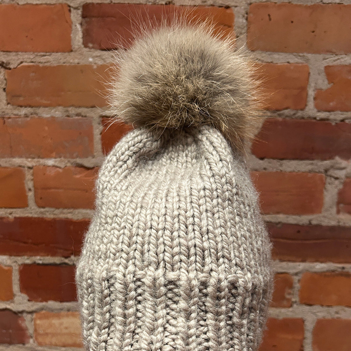 A light gray knit beanie with a large brown Coyote-fur pom-pom, displayed on a mannequin head with red lips in front of a red brick wall.
