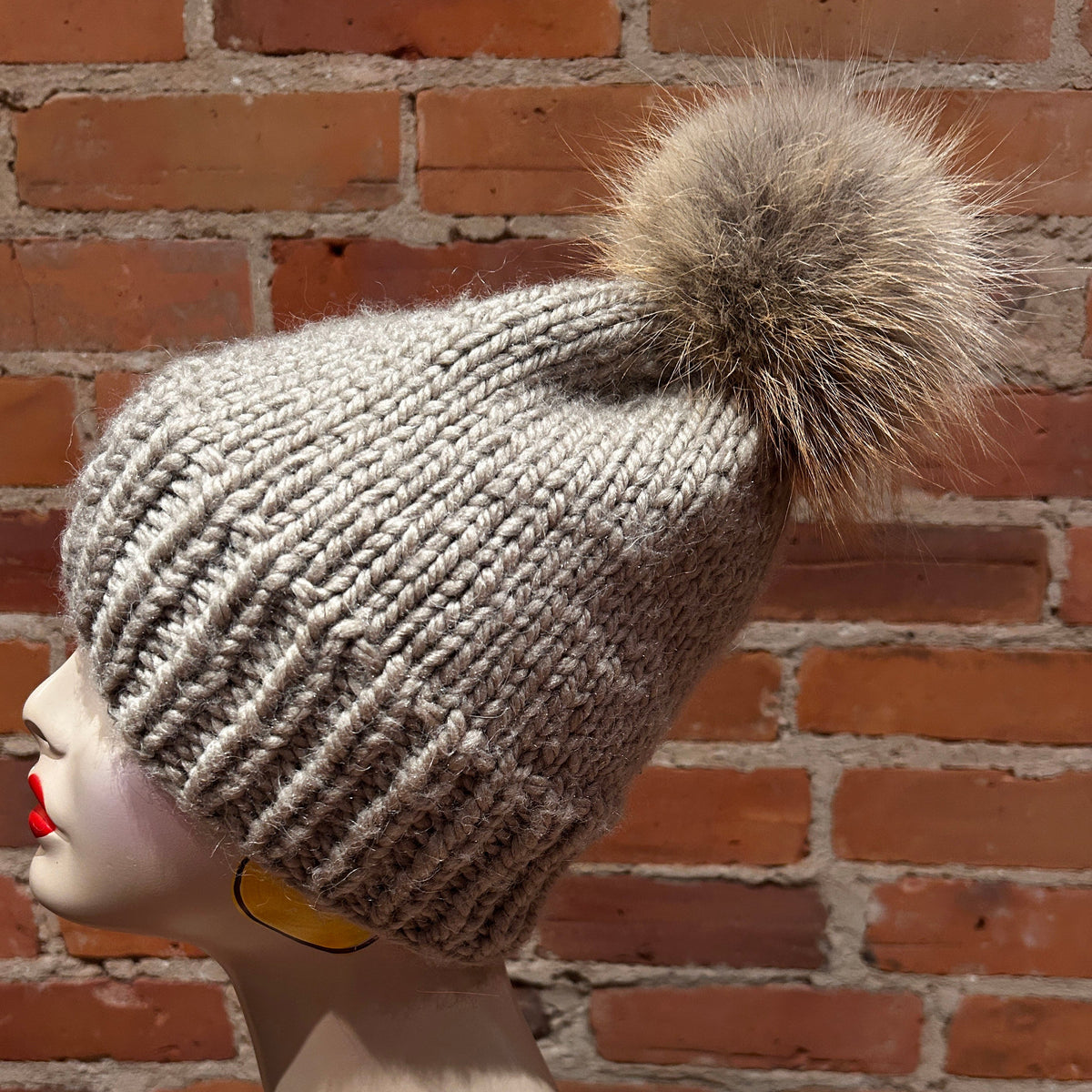 A chunky knit beanie hat with a large fluffy pom-pom, shown on a mannequin head in profile against a red brick wall background.