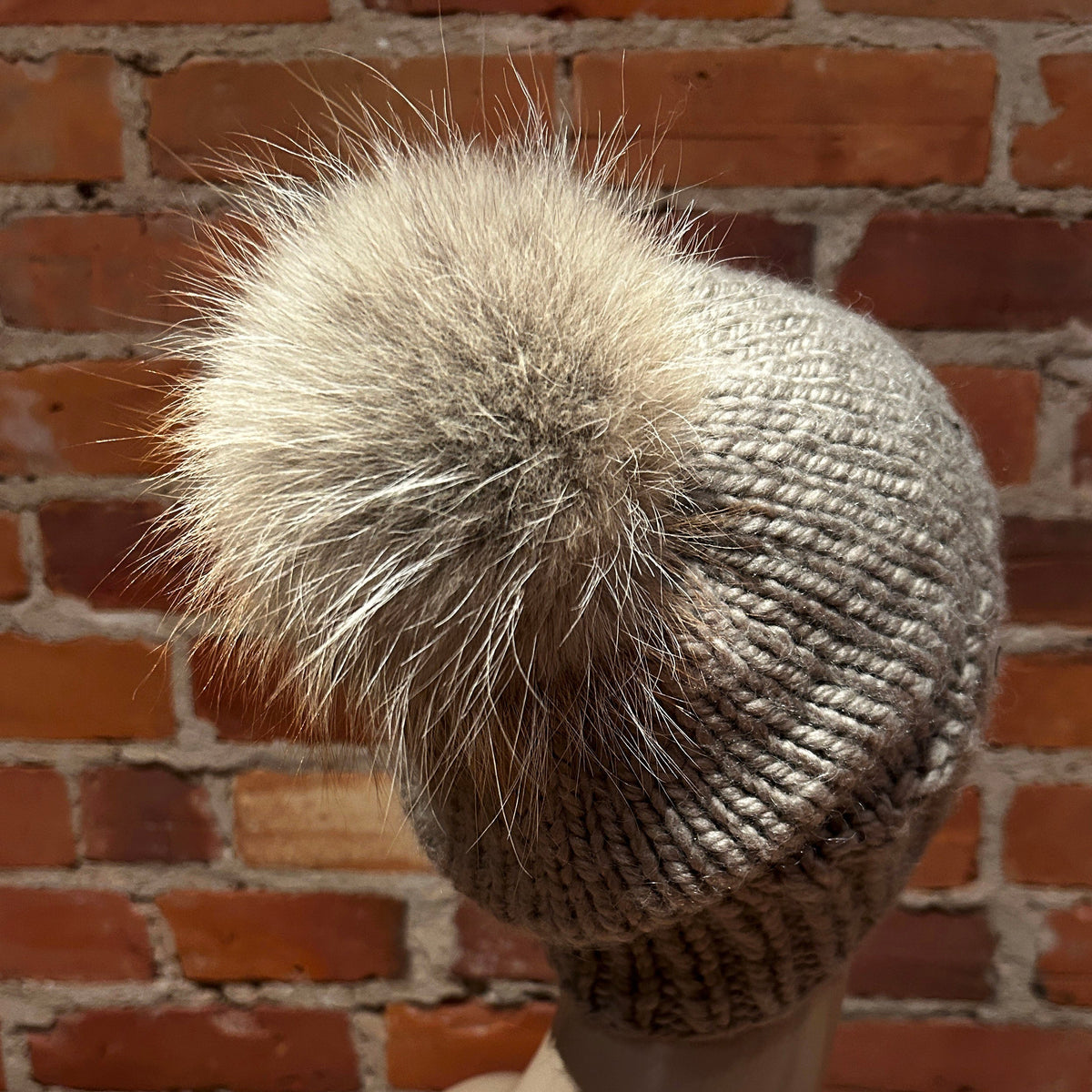 A close-up side view of a chunky knit beanie hat with a large fluffy pom-pom, displayed on a mannequin head against a red brick wall background.