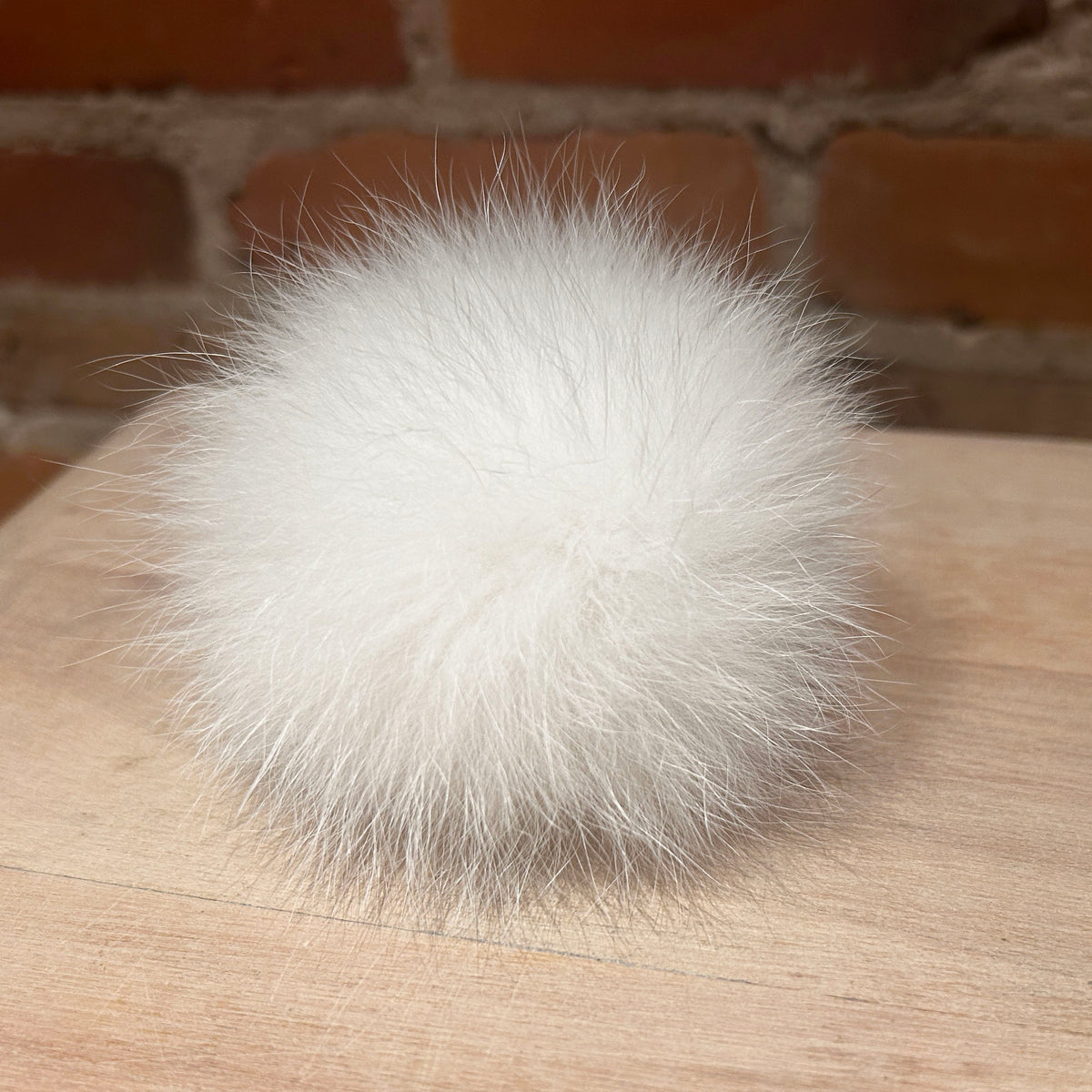 A fluffy white fox fur pom-pom ball resting on a light wood surface with a blurred brick wall background.