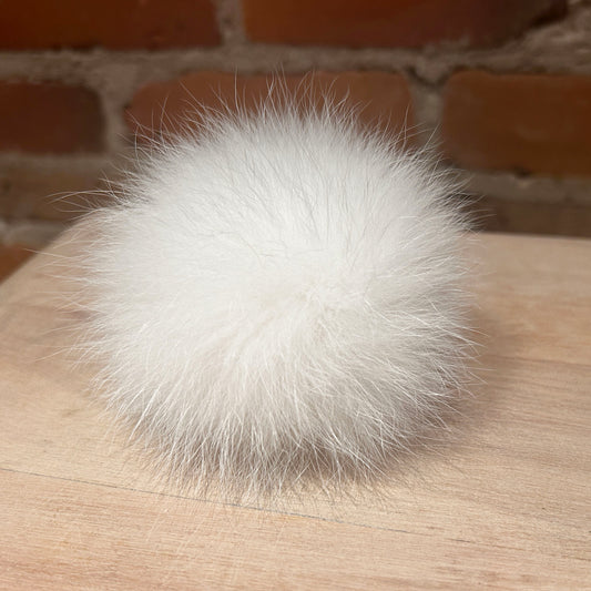 A fluffy white fox fur pom-pom ball resting on a light wood surface with a blurred brick wall background.