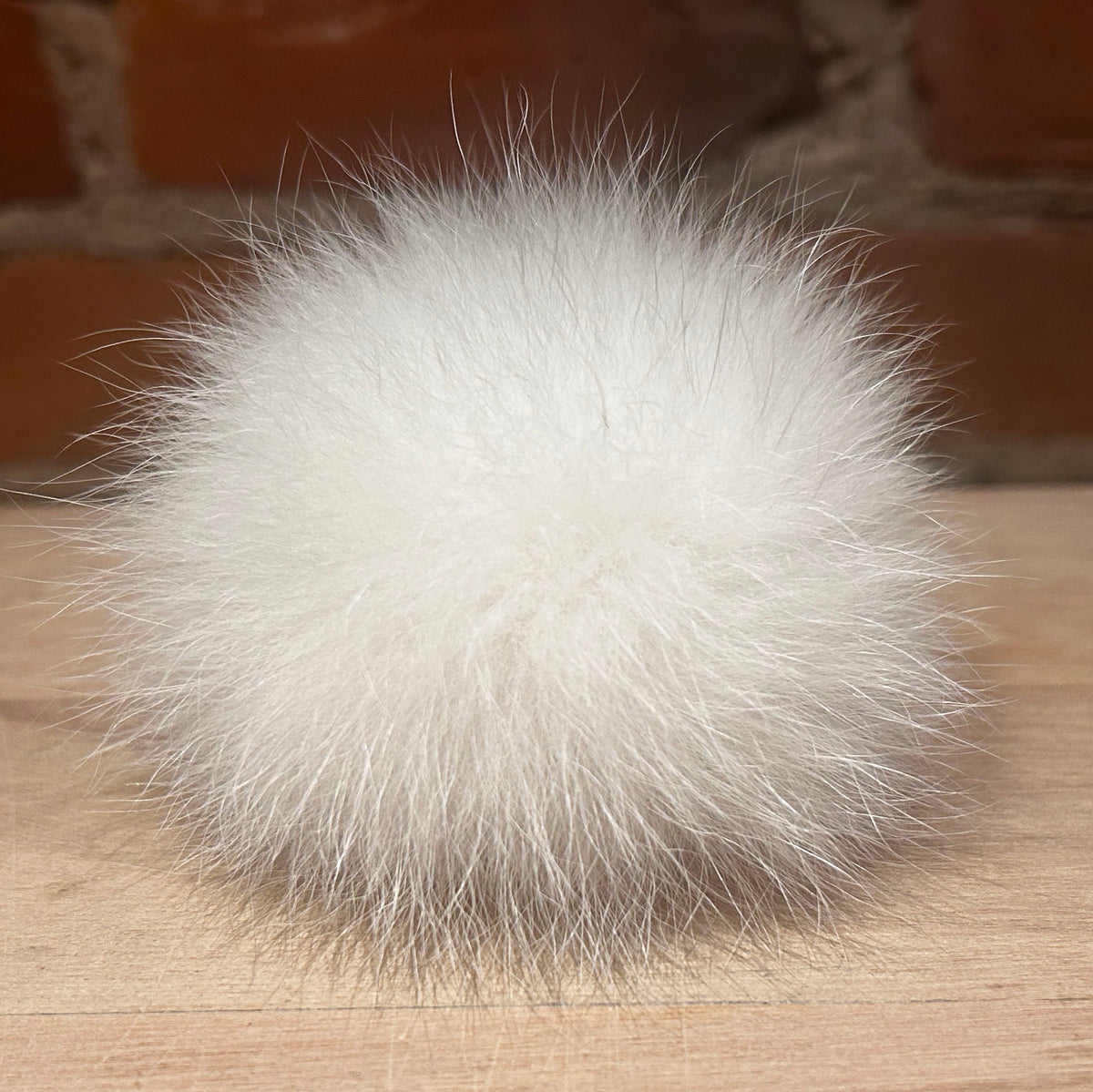 A fluffy white fox fur pom-pom ball resting on a light wood surface with a blurred brick wall background.