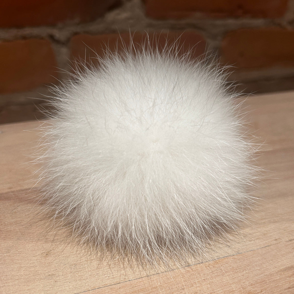 A fluffy white fox fur pom-pom ball resting on a light wood surface with a blurred brick wall background.