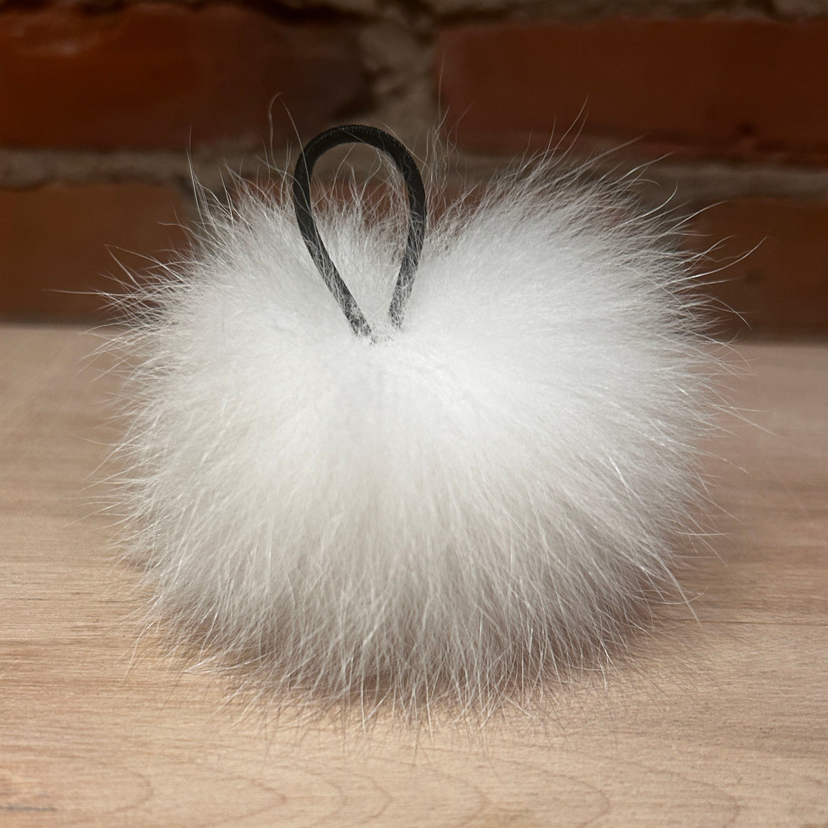 A fluffy white fox fur pom-pom keychain or bag charm with a black hanging loop, resting on a light wood surface with a blurred brick wall background.