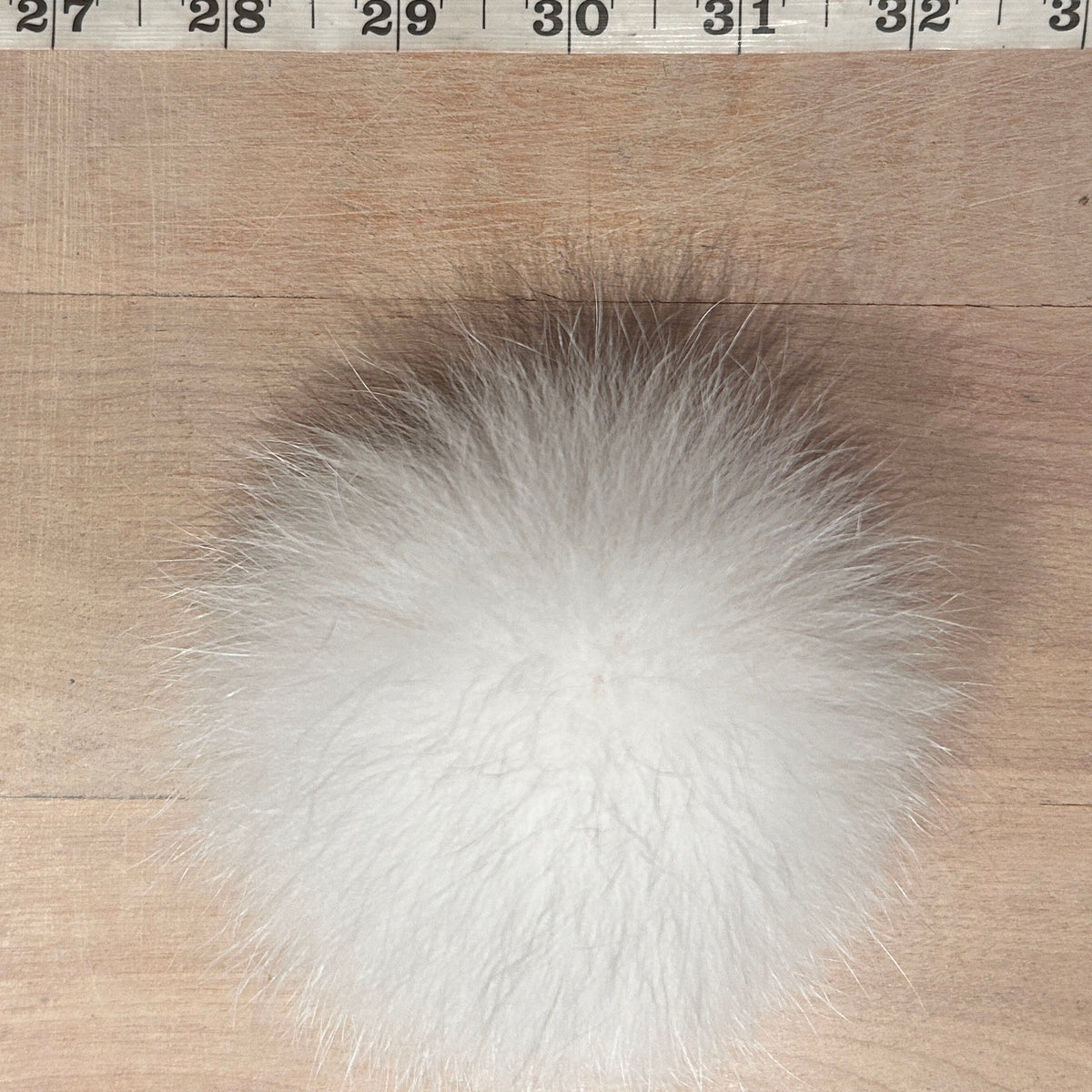 A fluffy white fox fur pom-pom ball shown on a light wood surface with a measuring tape ruler at the top for scale.