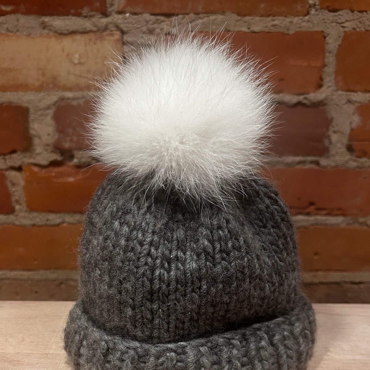 A small dark gray chunky knit beanie with a small fluffy white pom-pom, displayed on a light wood surface against a red brick wall background.