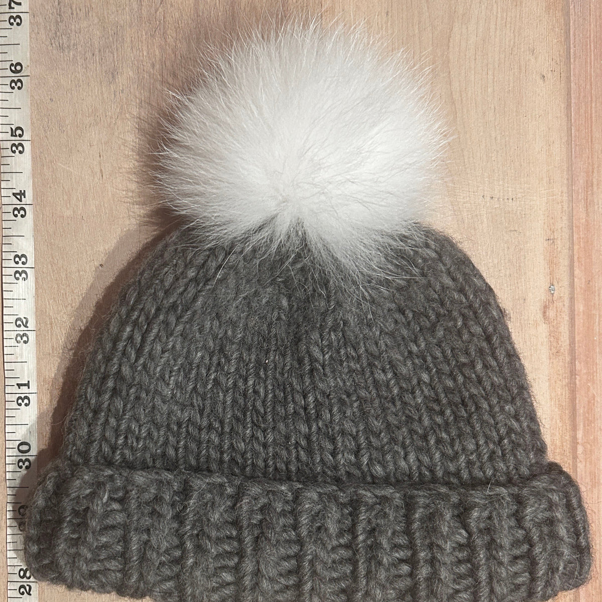 A small gray chunky knit beanie with a small white fox fur pom-pom, laid flat on a light wood background next to a measuring tape ruler.