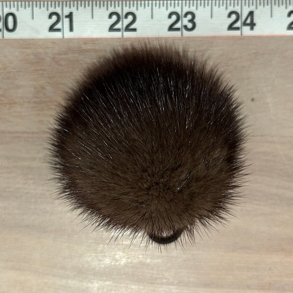 A dark brown fluffy pom-pom or hairball accessory shown from above on a light wood surface, with a measuring ruler visible along the top edge for scale.