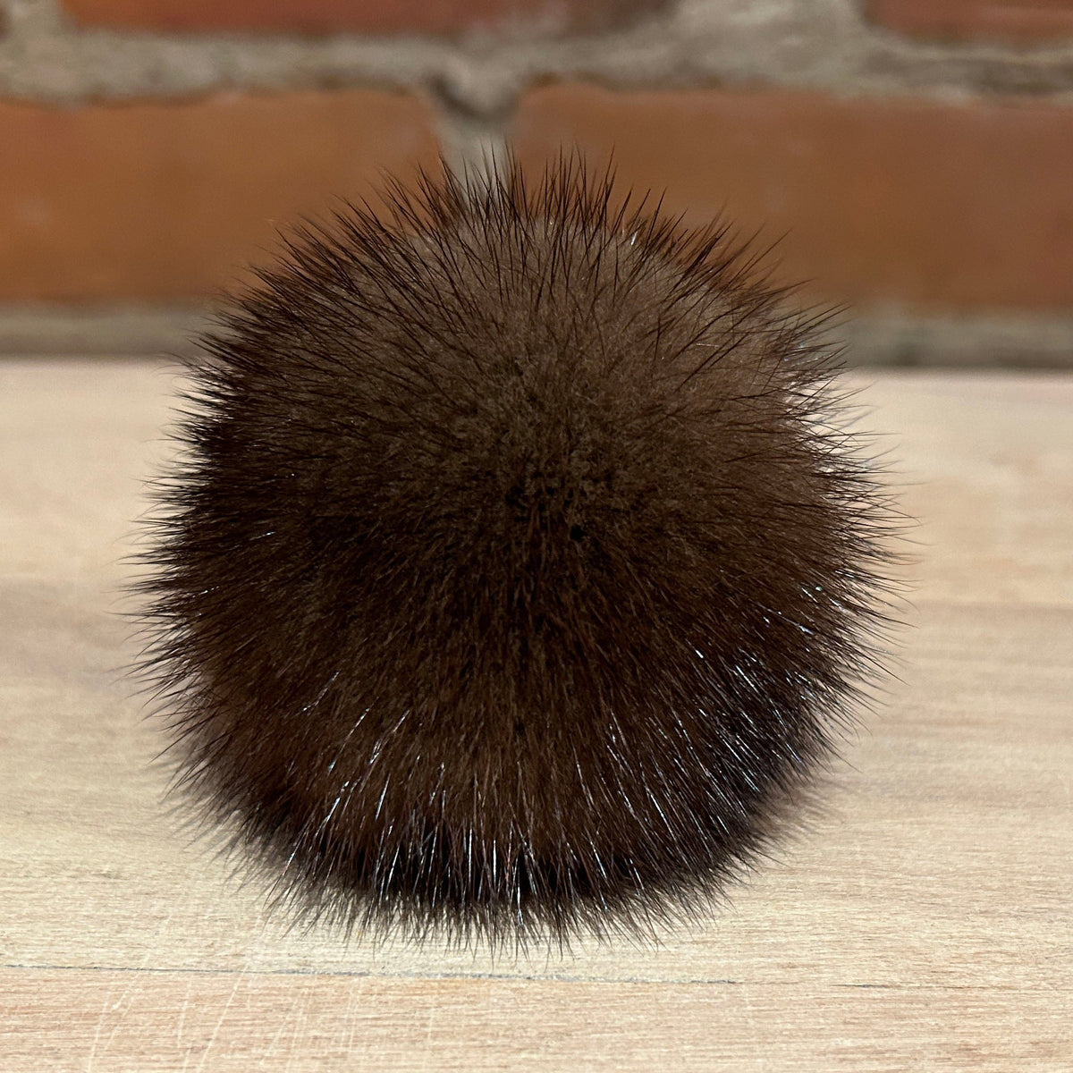 A dark brown, spiky pom-pom ball made of mink fur or synthetic fibers, resting on a light-colored wooden surface with a blurred brick wall in the background.