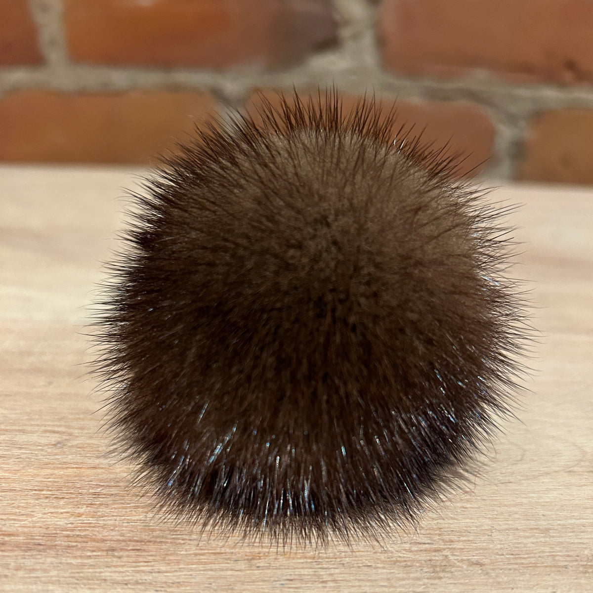 A round, dark brown mink fur pom-pom with spiky, bristly texture, resting on a light wood surface with a blurred brick wall background.