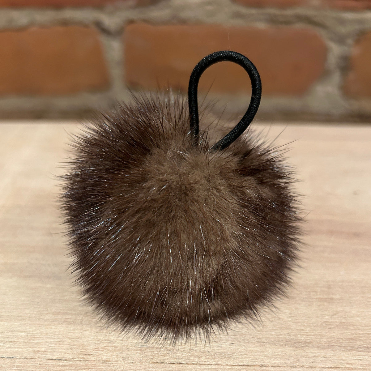 A brown mink fur pom-pom with a black elastic loop, resting on a light wood surface with a blurred brick wall background.