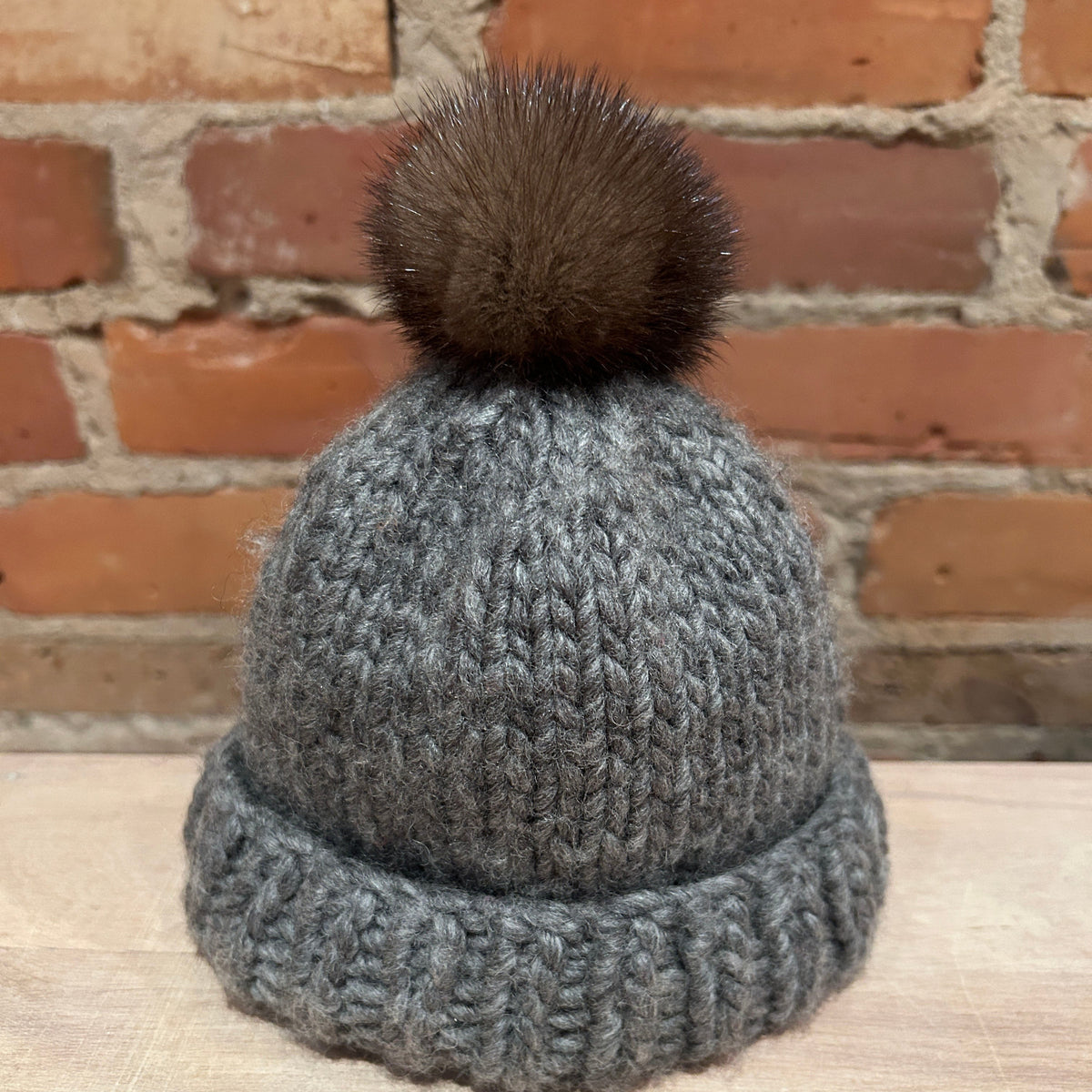 A small gray knit beanie hat with a small dark brown mink fur pom-pom, displayed on a light wooden surface against a red brick wall background.