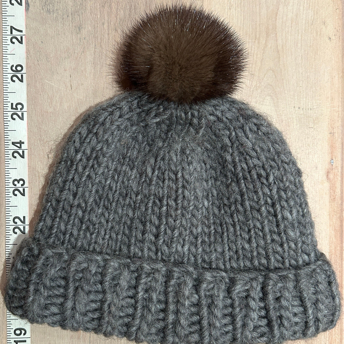 A gray chunky knit beanie with a brown mink fur pom-pom, laid flat on a light wood background next to a measuring tape.