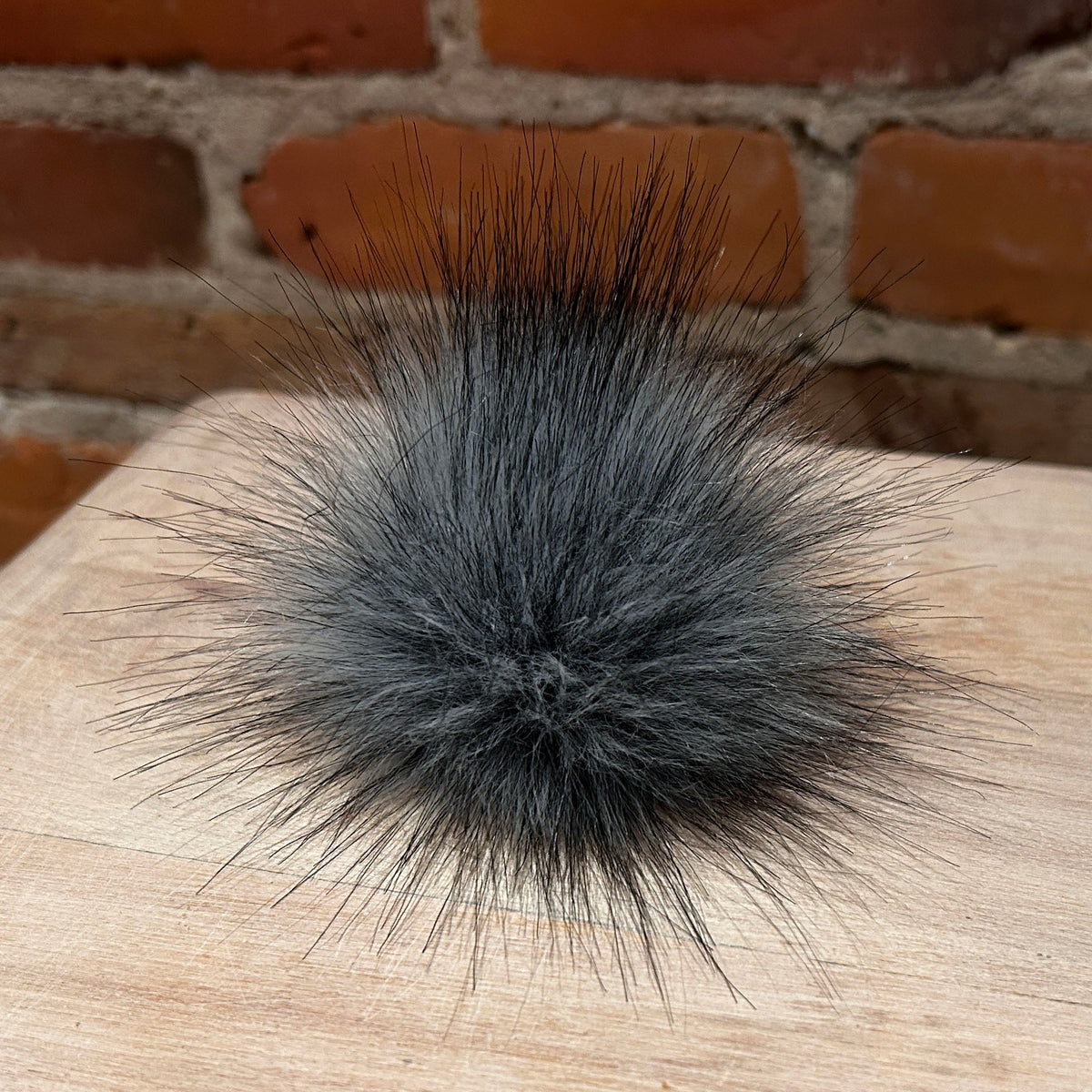 A fluffy dark gray faux fur pom-pom accessory resting on a light wood surface with a brick wall background.