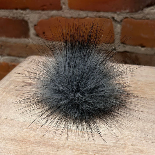 A fluffy dark gray faux fur pom-pom accessory resting on a light wood surface with a brick wall background.