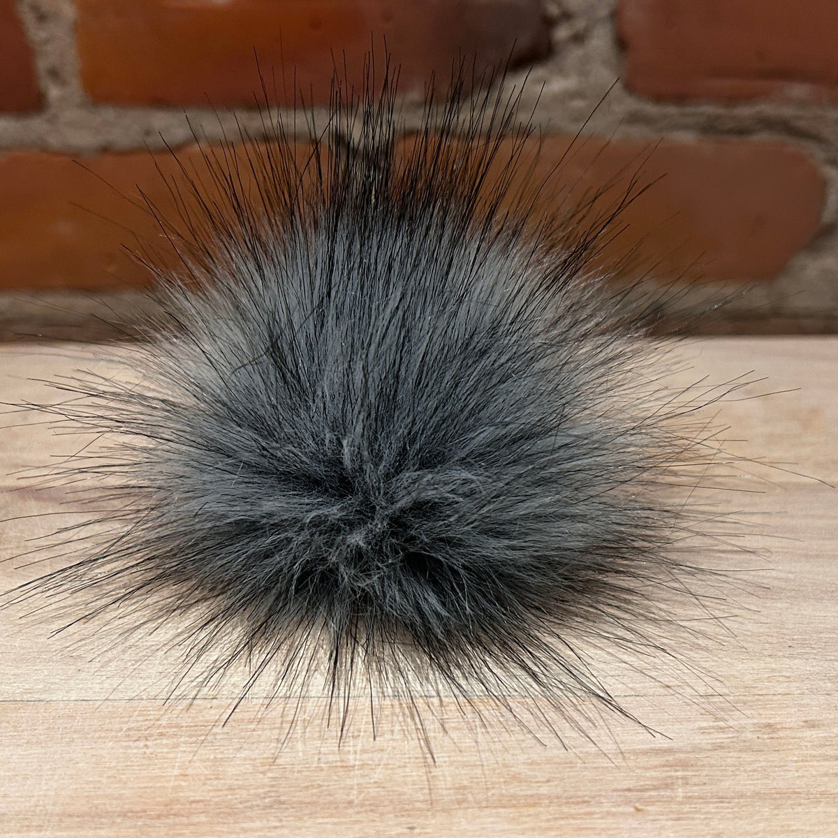 A fluffy gray faux fur pom-pom with spiky black and gray hairs, resting on a light wood surface with a brick wall background.