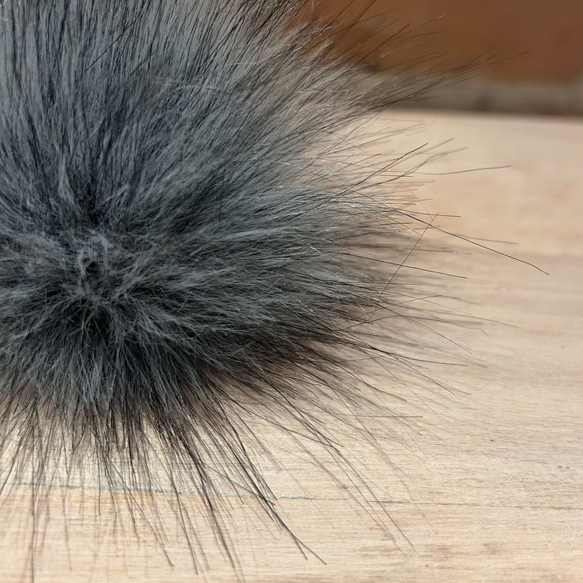 A close-up of a fluffy dark gray faux fur pom-pom with long, wispy fibers, resting on a light wood surface with a blurred brick wall background.