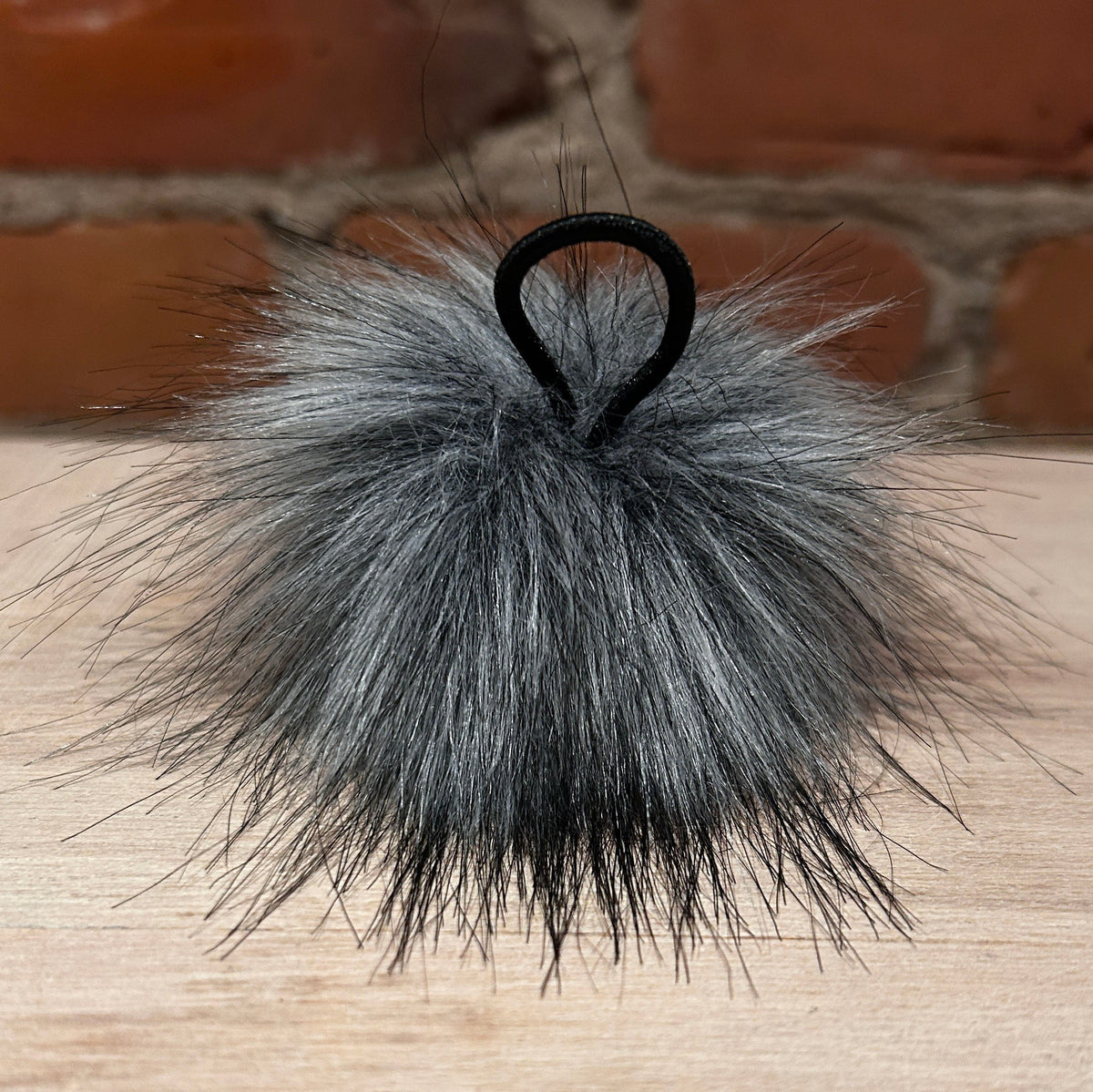 A gray faux fur pom-pom hair accessory with a black elastic loop, resting on a light wood surface with a brick wall background.